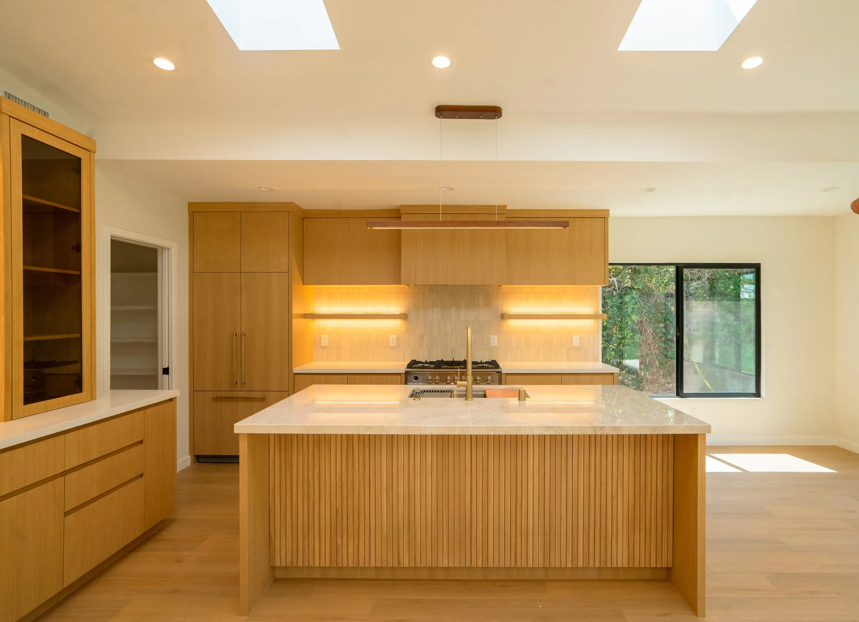 Oak kitchen with reeded island, pale stone counters, illuminated shelves, and skylights