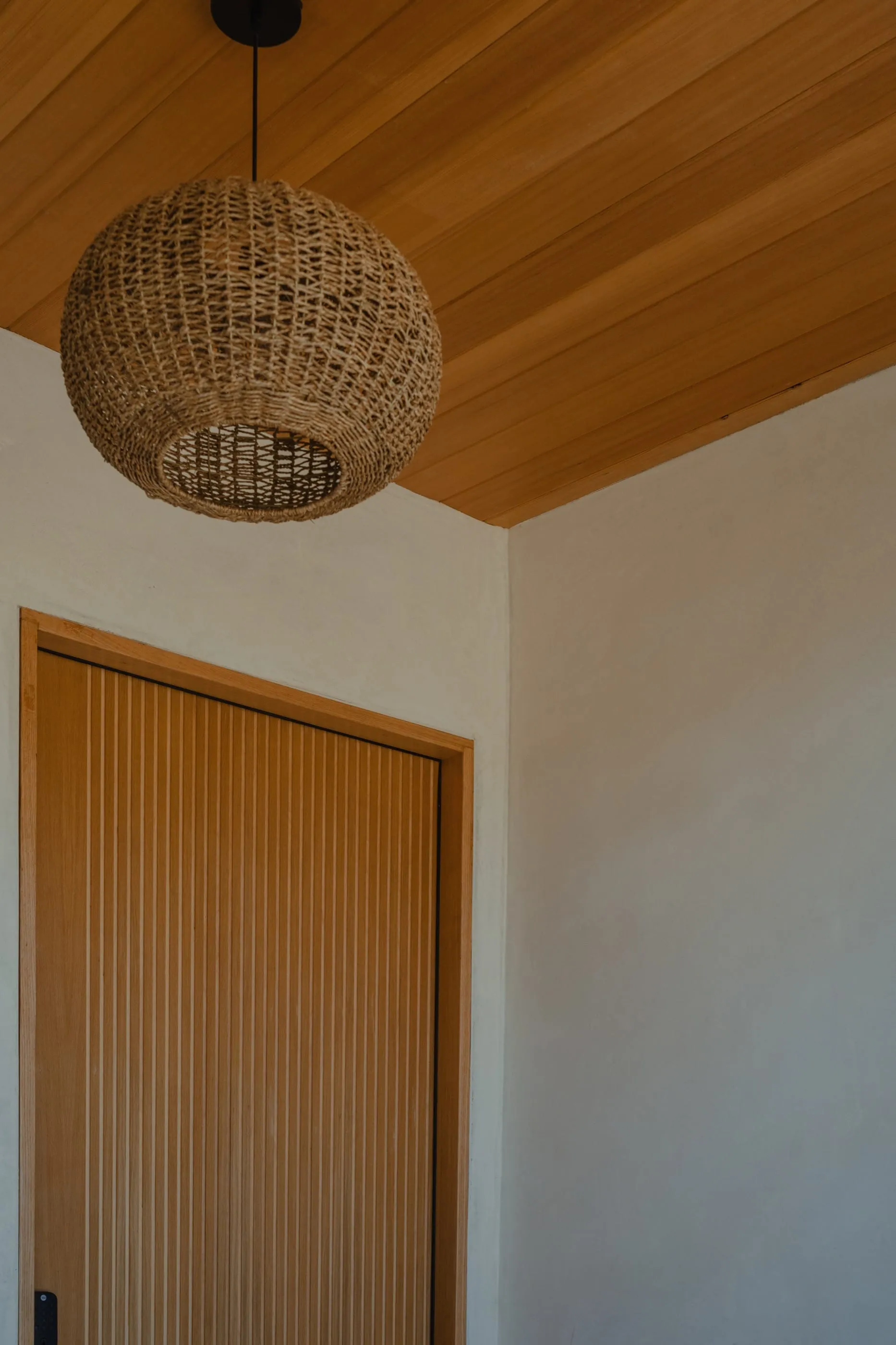 Slatted oak entry door beneath a woven pendant and wood ceiling