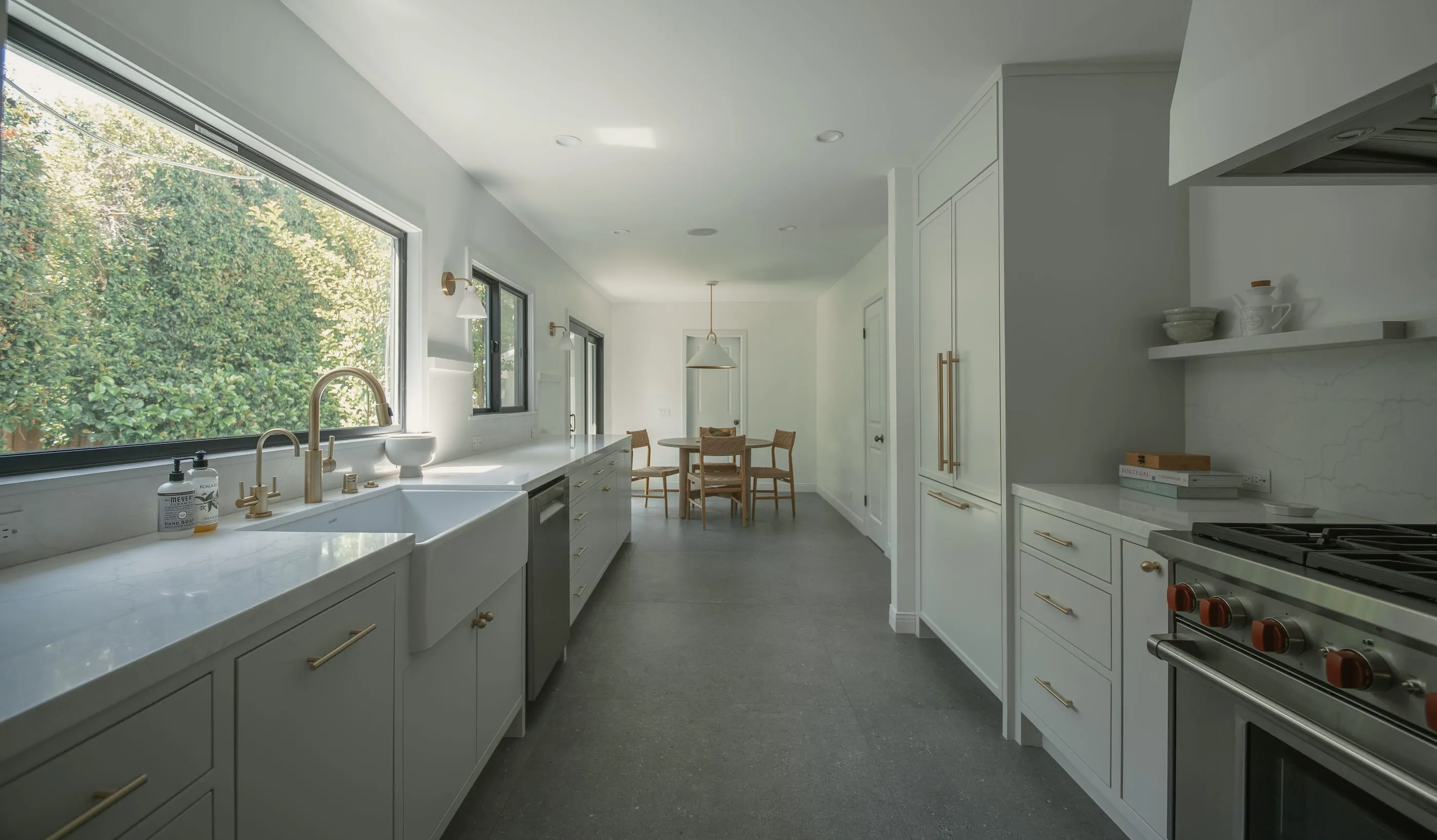 Long view through a pale kitchen with stone counters, brass hardware, and a breakfast table