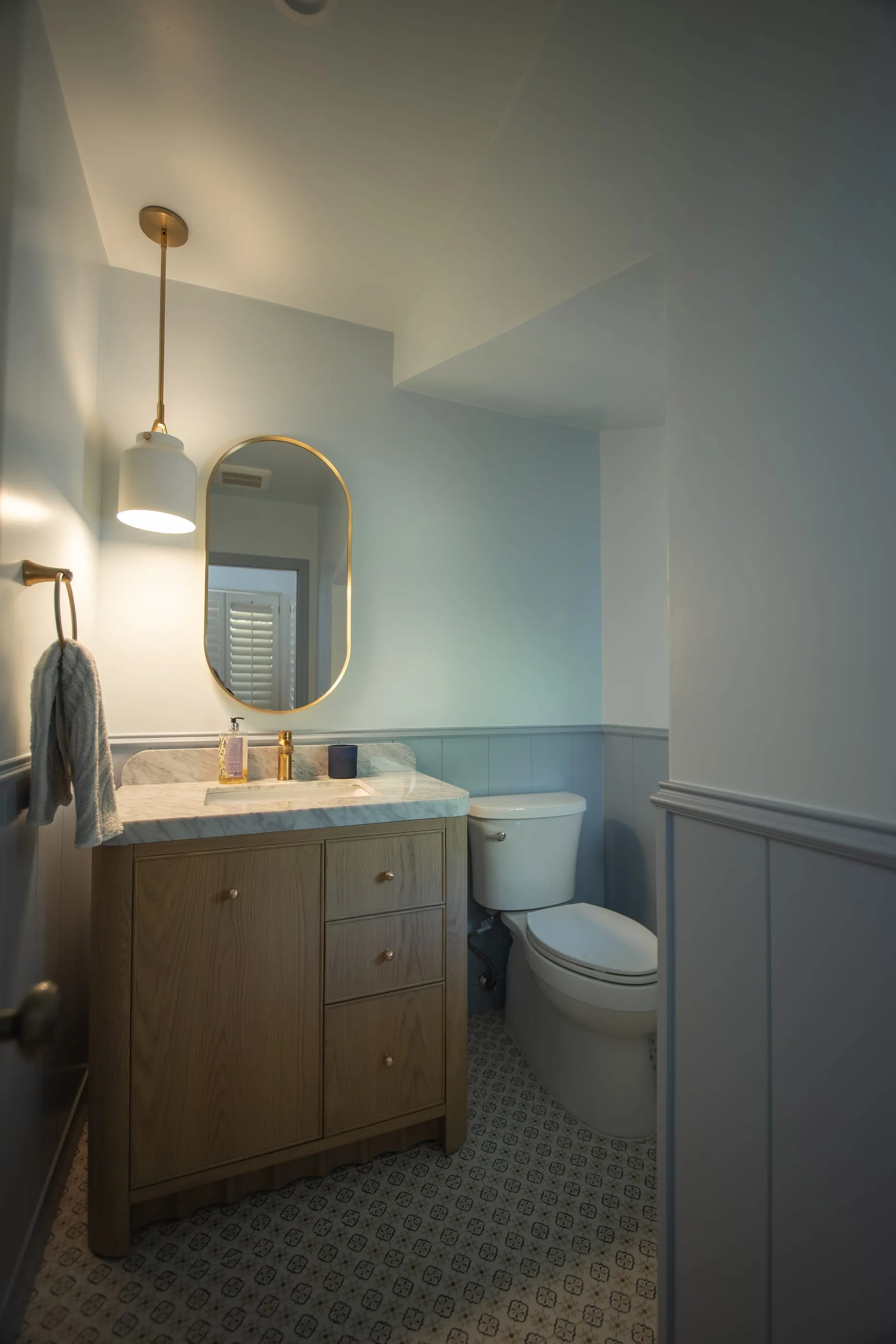 Powder room with an oak vanity, marble counter, blue-gray paneling, and brass pendant