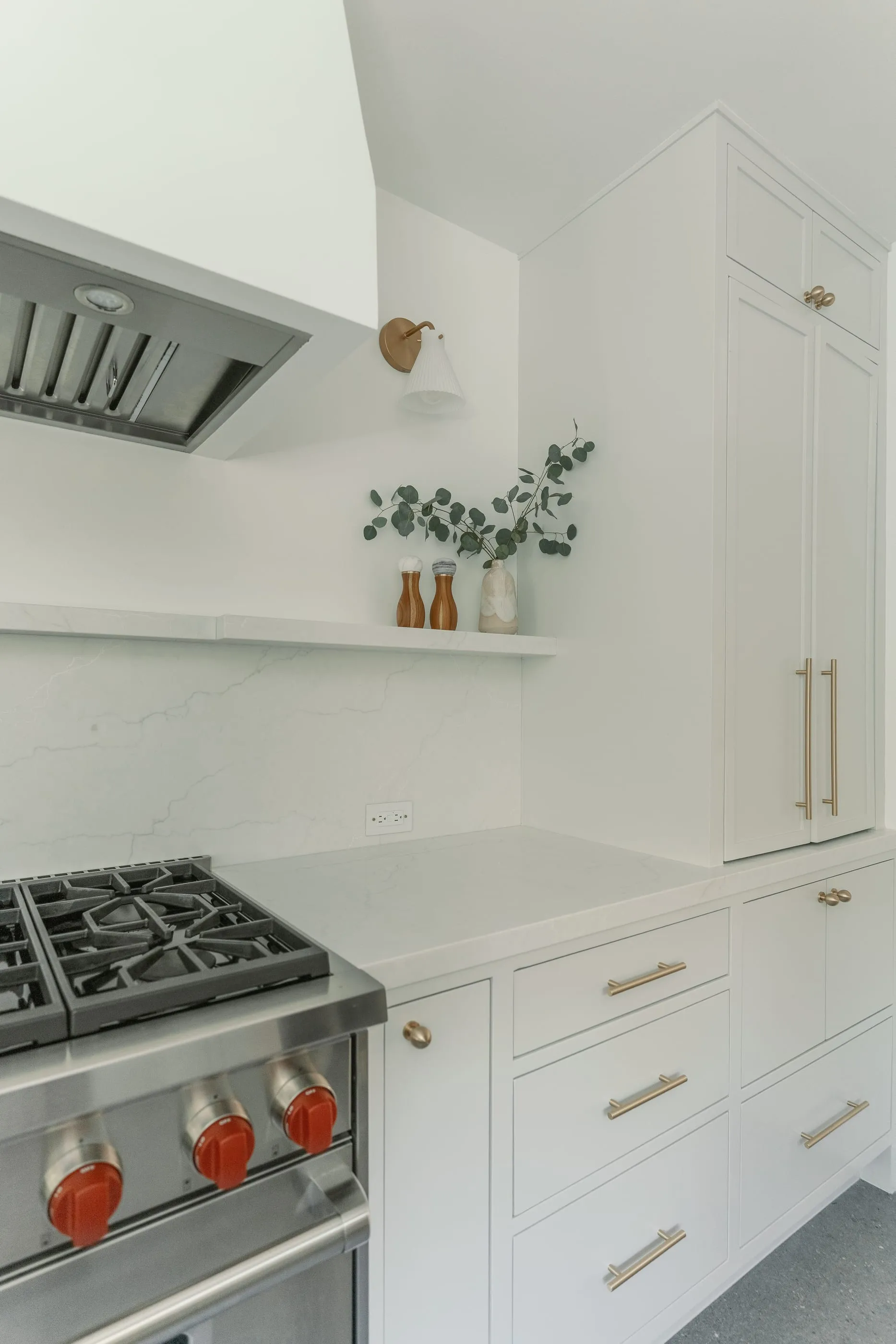 Range wall with pale cabinetry, stone backsplash, brass hardware, and a narrow display shelf